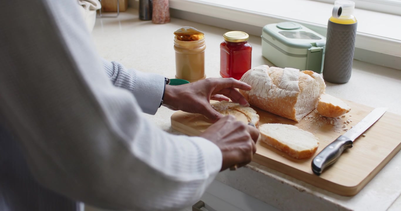 Uomo afroamericano con un maglione prende un coltello, raccoglie la marmellata e la spalma sul pane per mangiare al bancone.