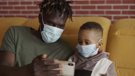 padre e hijo están sentados en un sofá con máscaras médicas, viendo video en un teléfono inteligente
