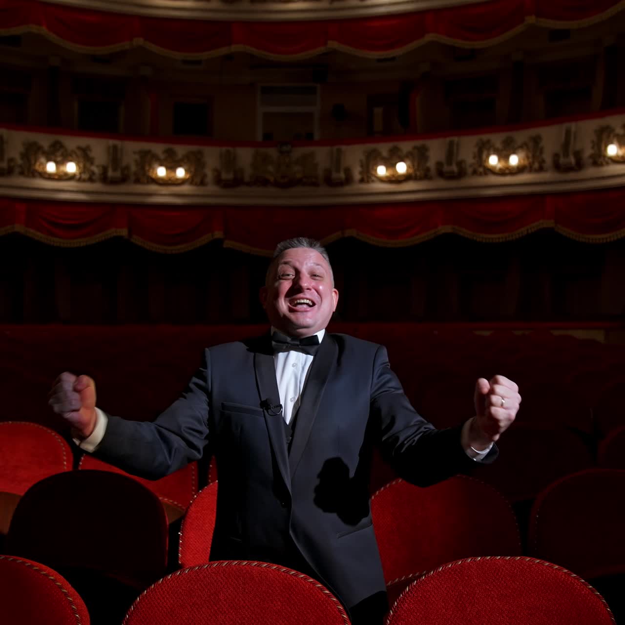 Happy actor in dark theatrical hall. Talented man in suit standing among vacant red chairs and shouting cheerfully. Portrait of a comedian in empty theater