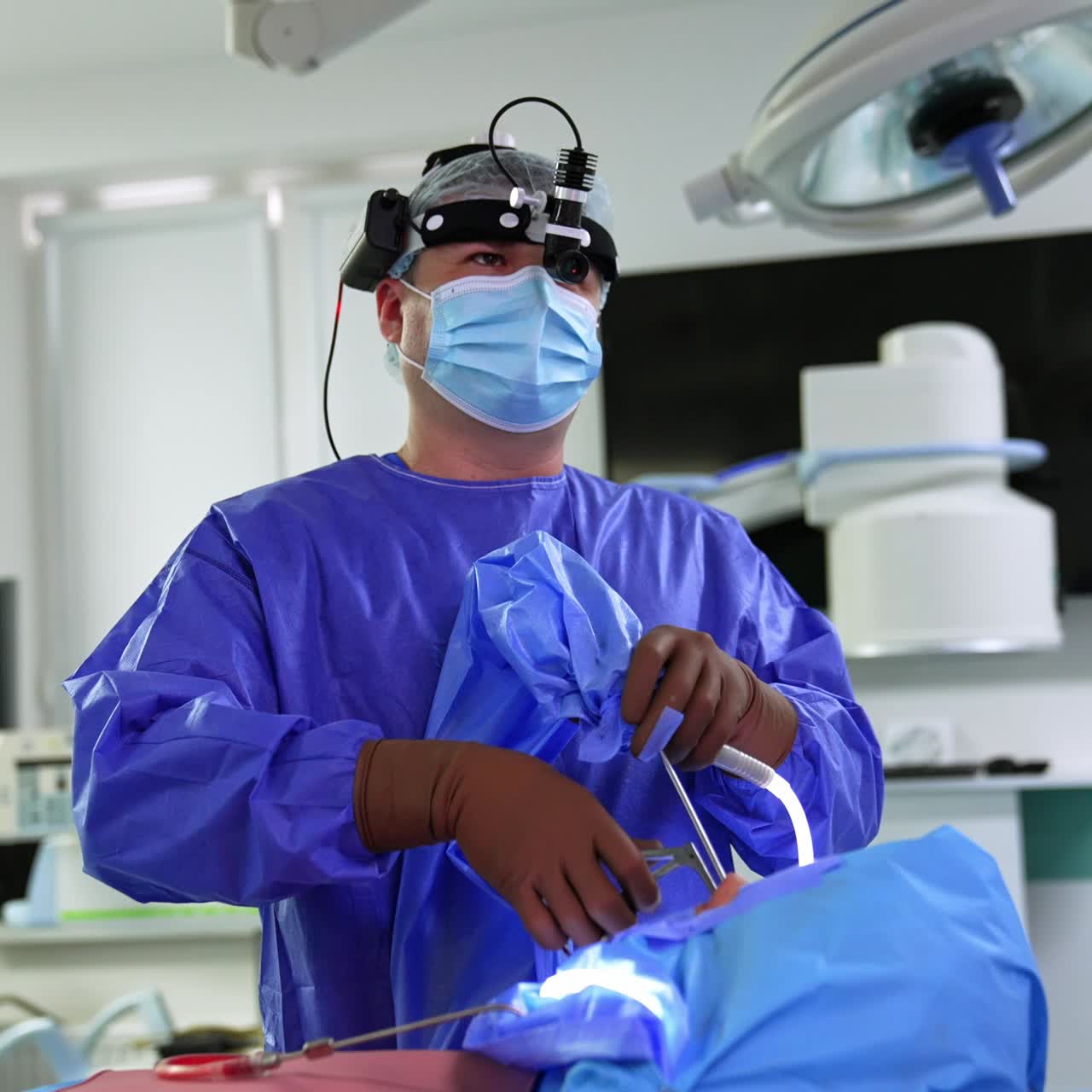 Confident otolaryngology surgeon quickly moving tool in patient's nose. Doctor with headlight looks at the screen in front of him intently. Female nurse assisting