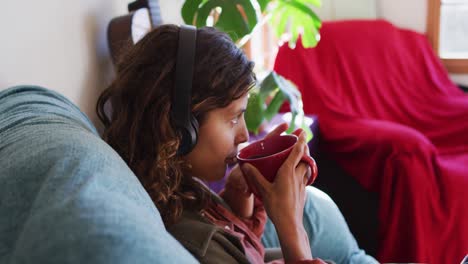 Relaxed-mixed-race-woman-wearing-headphones-sitting-drinking-tea-in-sunny-cottage-living-room