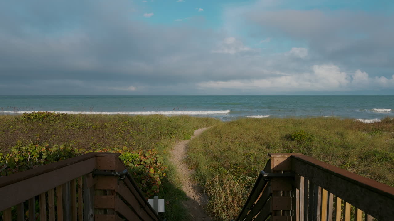 A wooden boardwalk ends at a beach access path, framed by grassy dunes and a surfside showerhead, with the ocean stretching out beneath scattered clouds