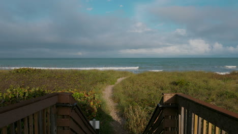 a wooden boardwalk ends at a beach access path, framed by grassy dunes and a surfside showerhead, with the ocean stretching out beneath scattered clouds