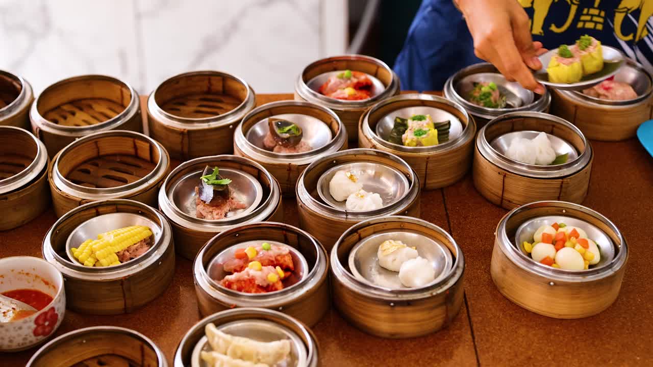 Hands arrange colorful dim sum in bamboo steamers on a table under bright lighting, capturing a vibrant culinary scene