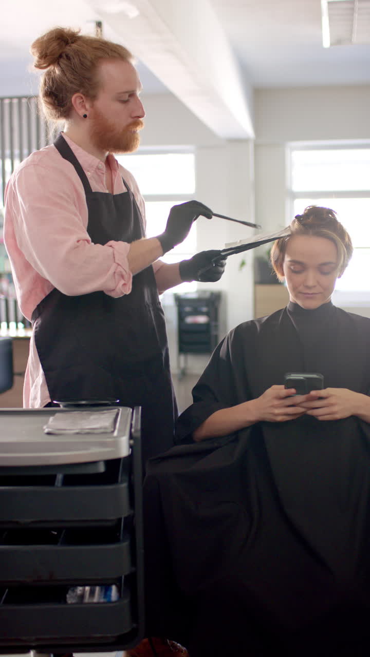 Verticale video van een Kaukasische mannelijke kapper die het haar van een klant verft in een kapsalon, in slow motion.