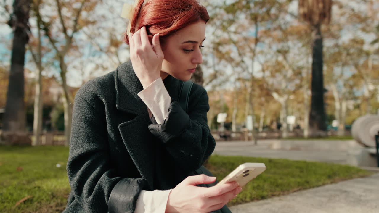 Рыжеволосая молодая женщина использует смартфон и беспроводные наушники в парке