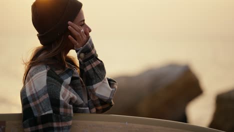 A-happy-blonde-girl-in-a-black-hat-and-a-plaid-shirt-stands-near-a-rocky-shore,-holds-a-surfboard-and-looks-towards-the-sea-in-the-morning