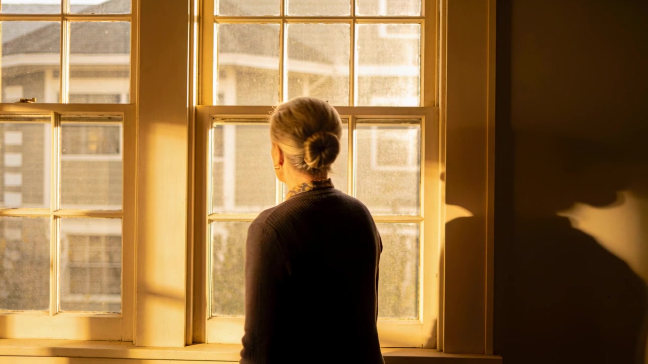 Streaming sunlight nudging senior woman in cardigan turning at tall paned window, casting shadow