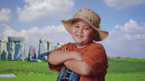asian boy with a hat and binoculars crossing arms and smiling to camera while traveling in stonehenge. boy researcher examines something, travel adventure, close up side view