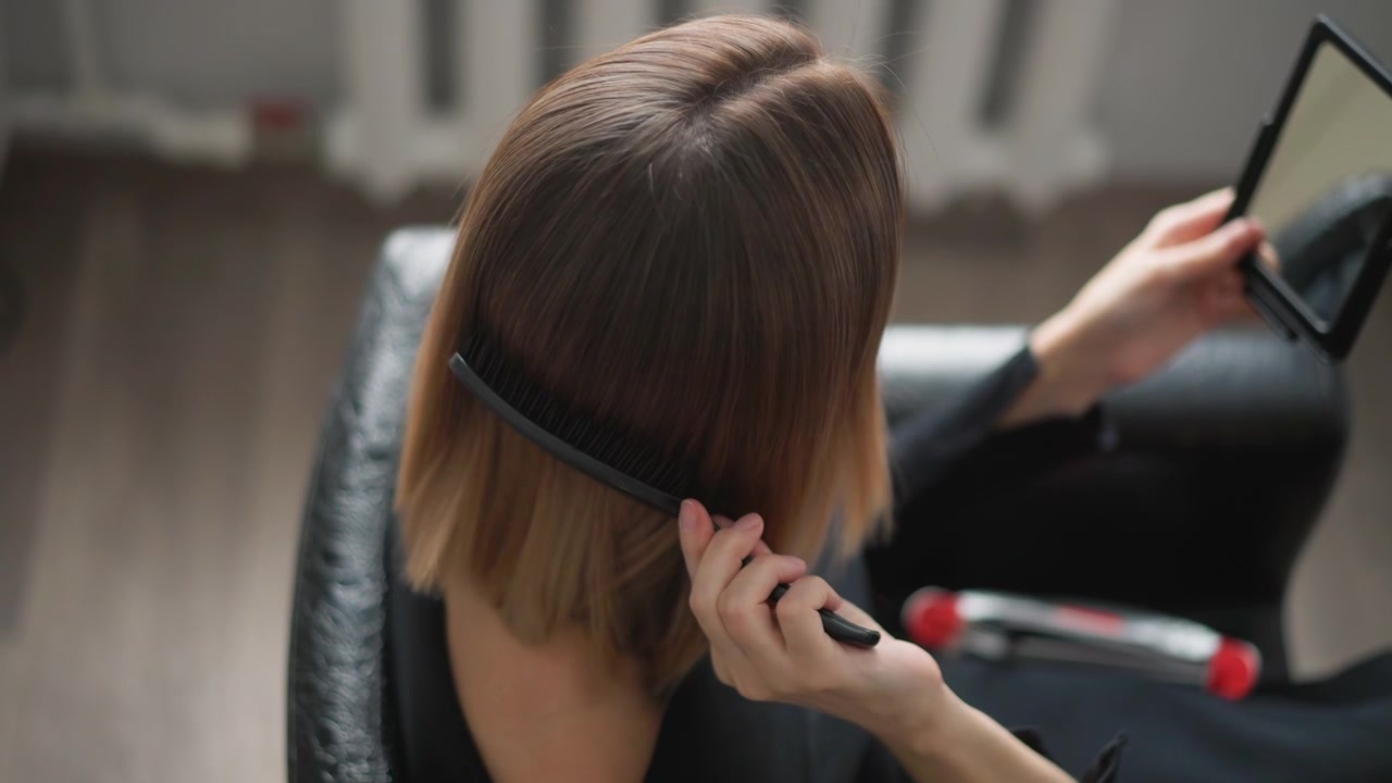 Primer plano de una mujer cepillándose y peinándose el cabello mientras se mira en un espejo de mano. La mujer usa varios cepillos para alisar los mechones, con una iluminación suave que resalta el detalle de la textura en interiores.