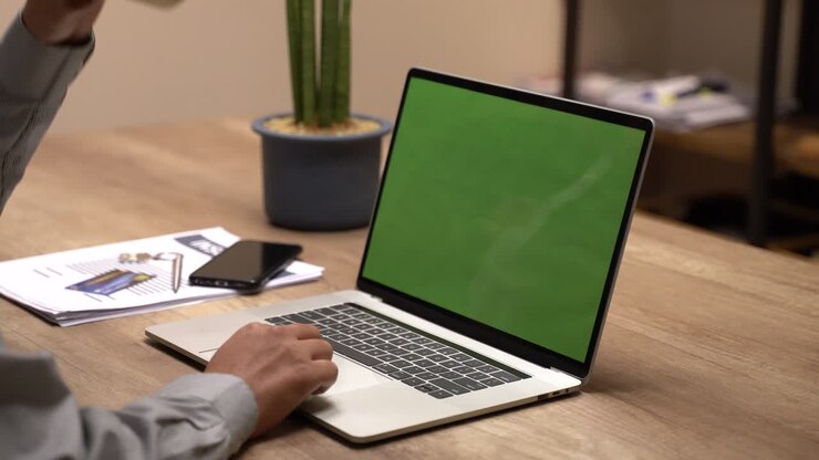 Nahaufnahme der Hände eines Mannes, der an einem Laptop mit Greenscreen arbeitet. Im Hintergrund ein unscharfes und hell beleuchtetes Büro, wo ein Mann auf den anderen zugeht und sie sich unterhalten.