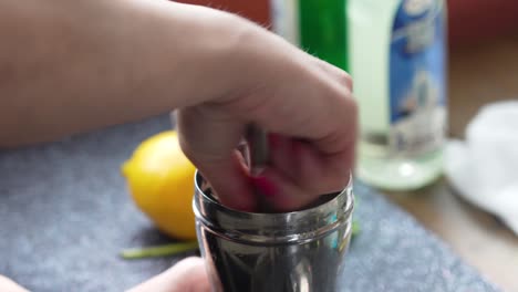 Bartender-Twists-and-Muddles-in-a-Silver-Cobbler-Shaker-with-a-Muddler-to-make-a-Cocktail-or-Mixed-Drink-with-Citrus-and-Mint-Leaves,-Closeup-of-Beverage-and-Woman’s-Hands-with-Lemon-in-Background