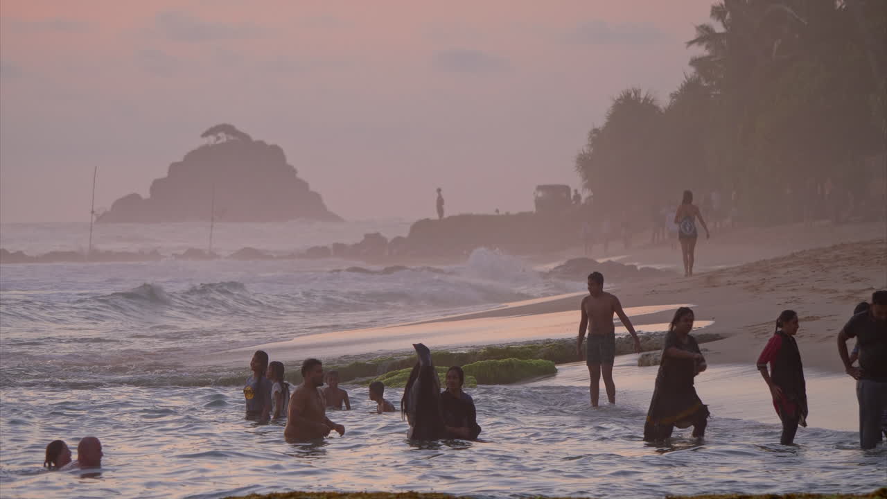People splash in the ocean at sunset. Children jump into the water. Waves churn on the shore. The sky is dyed pink
