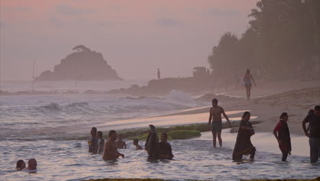 people splash in the ocean at sunset. children jump into the water. waves churn on the shore. the sky is dyed pink