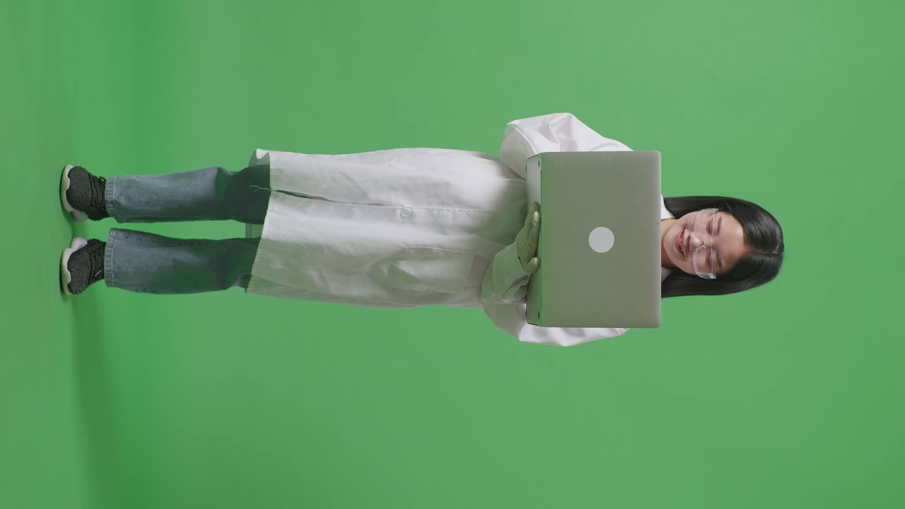 cuerpo lleno de una mujer científica asiática sonriente trabajando en una computadora portátil mientras está de pie en el fondo de la pantalla verde en el laboratorio