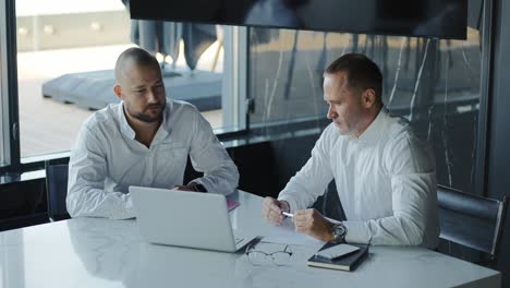 Two-diverse-businessmen-discussing-financial-market-data-using-laptop-and-digital-tablet.-Financial-advisor-broker-manager