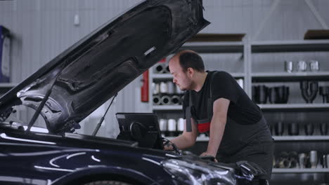 man mechanic at the hood of a car with a tablet diagnoses all systems and searches for malfunctions