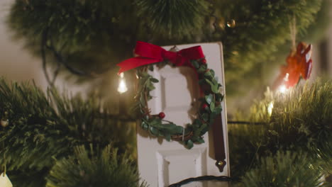 Camera-Focuses-On-White-Door-Decorated-With-Christmas-Wreath-Hanging-On-Christmas-Tree-With-Lights-1