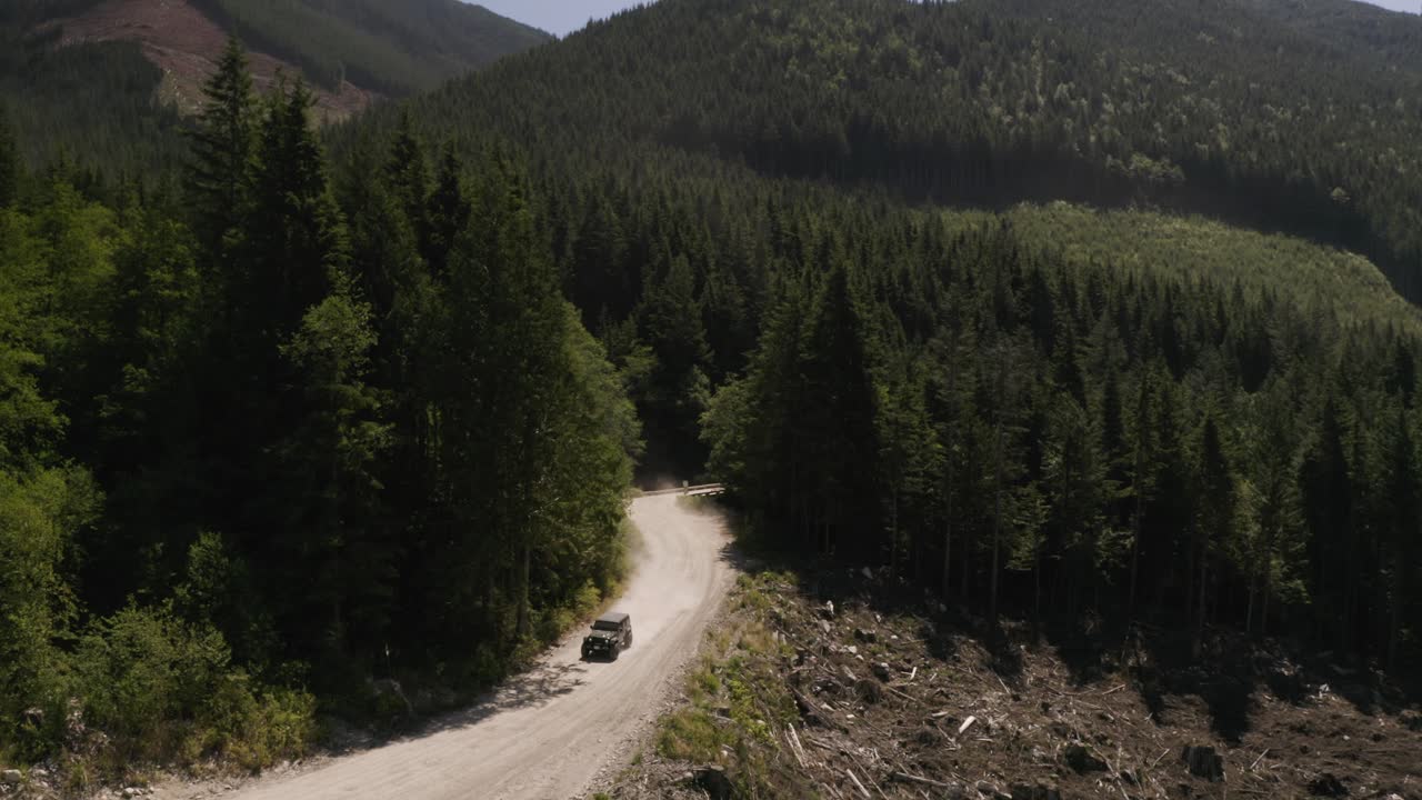 panorámica aérea a la izquierda de un jeep conduciendo por una polvorienta carretera en la ladera rodeada por un denso bosque de pinos verdes, montañas en el fondo, columbia británica, canadá