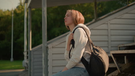 young woman in casual attire stands beside stadium bleachers, adjusting her black bag strap with a focused expression as she sit down on the bleacher