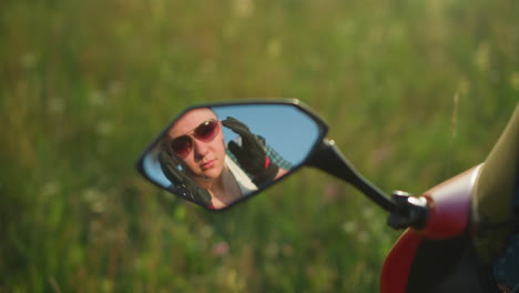 reflejo en el espejo de una mujer ajustando sus gafas de sol mientras usa guantes, con un campo de hierba borroso en el fondo, parece enfocada, con la luz del sol iluminando suavemente su cara