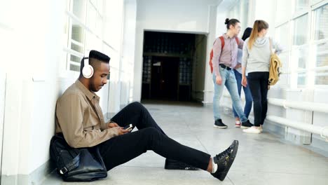 young handsome african american student sitting on floor in white corridor with headphones on head listening to music holding smartphone texting someone. his classmates are behind him