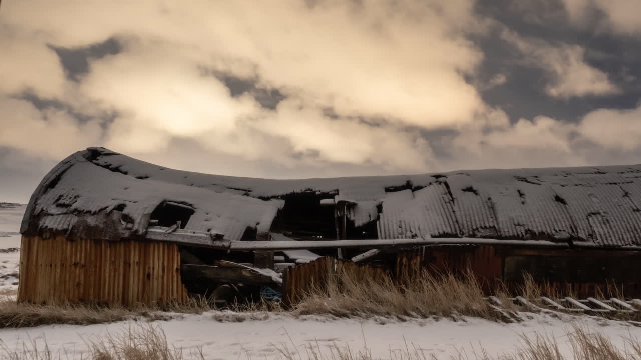 12 second stable timelapse overlooking an abandoned, broken barn covered in snow. Clouds lit up by city lights move across the shot.