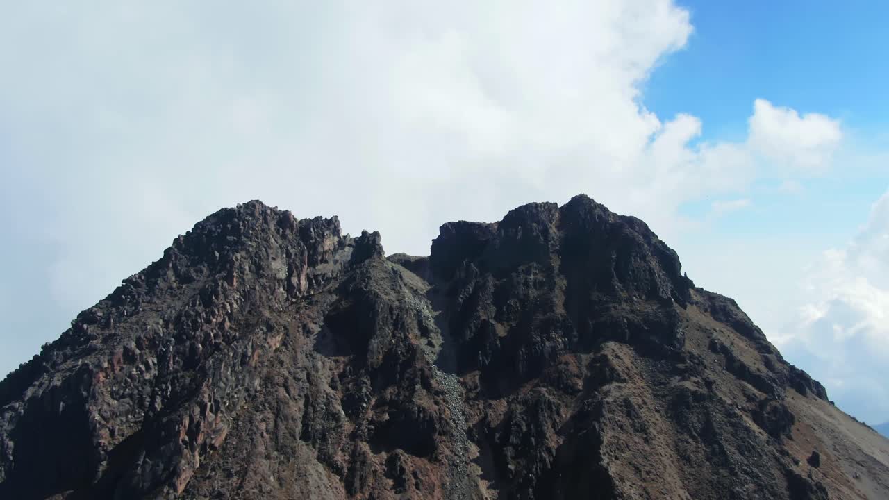 Aerial dolly in shot approaching the rocky summit of Nevado de Colima (Zapotepetl), with steep volcanic slopes and clouded sky in the background