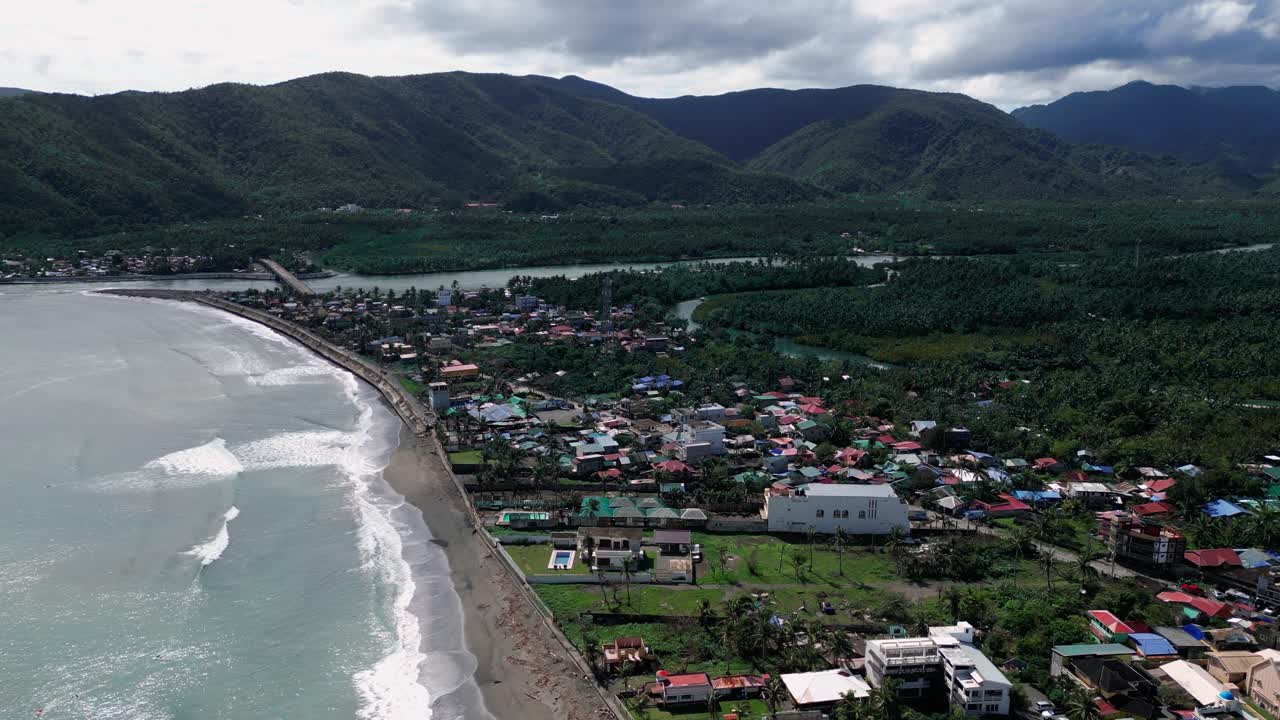 A wide shot showcases mountains in the background, a clear sky above, trees lining the shore, and beach houses along the coastline, creating a serene coastal scene where the ocean meets the land.