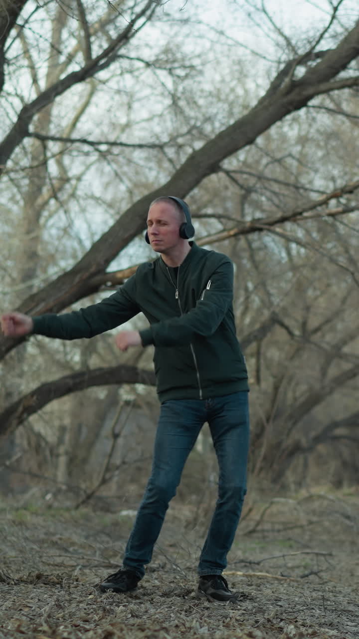 un hombre con una chaqueta verde con auriculares baila solo en un bosque sereno, el suelo está cubierto de hojas y ramas