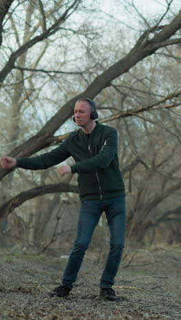 a man in a green jacket with headphones dances alone in a serene forest, the ground is covered with leaves and twigs