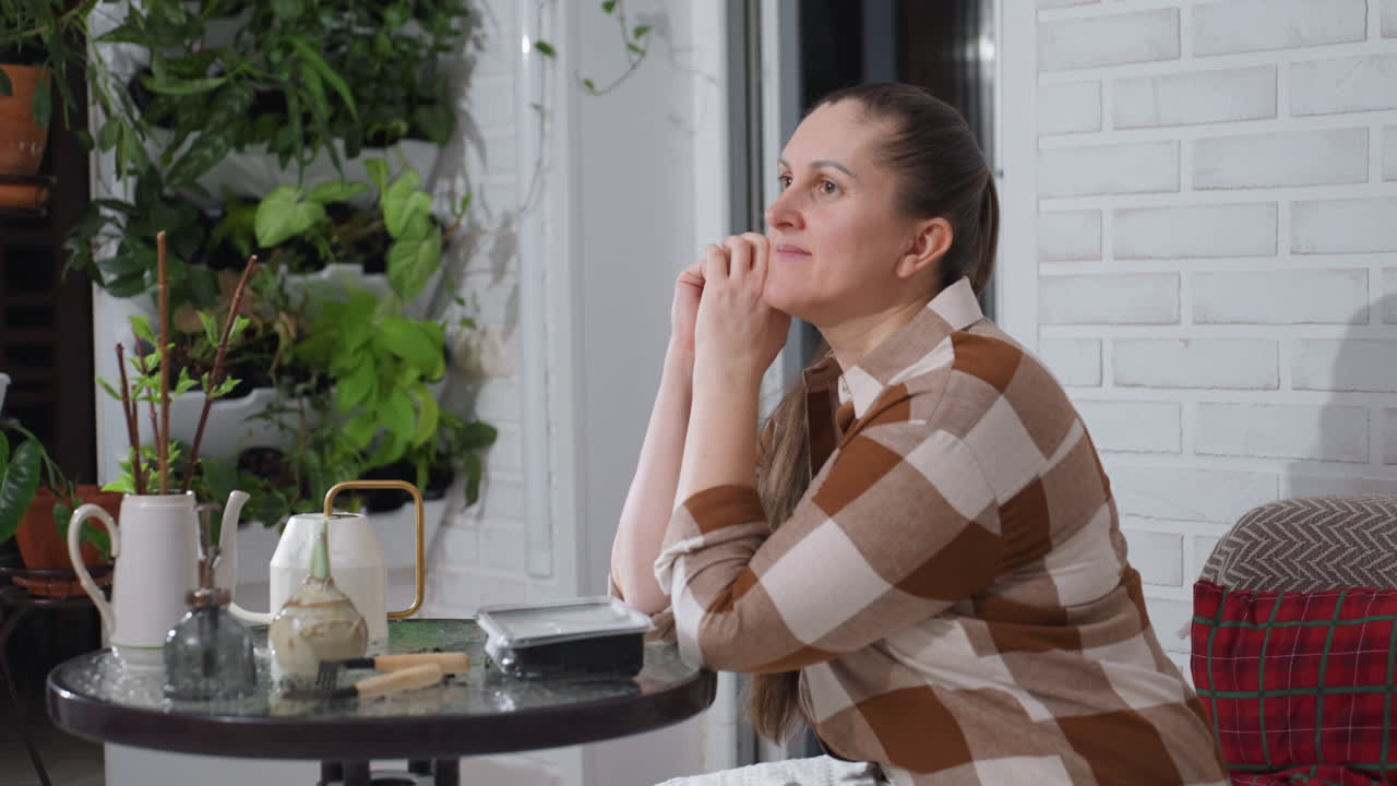 Portrait of woman leaning on hands at glass table surrounded by indoor plants against brick wall backdrop, gazing thoughtfully into distance with calm expression while gently supporting head on table