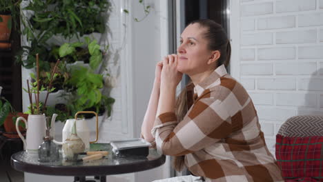 portrait of woman leaning on hands at glass table surrounded by indoor plants against brick wall backdrop, gazing thoughtfully into distance with calm expression while gently supporting head on table