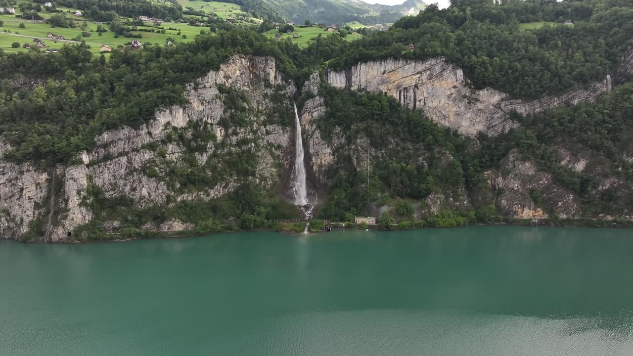A majestic natural scene captures the Seerenbach Falls cascading down steep, forested cliffs into the turquoise waters of Walensee in Switzerland. The view encompasses the dramatic alpine landscape