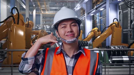 close up of asian male engineer with safety helmet smiling to camera and making call me gesture while standing with industrial robotic arms working
