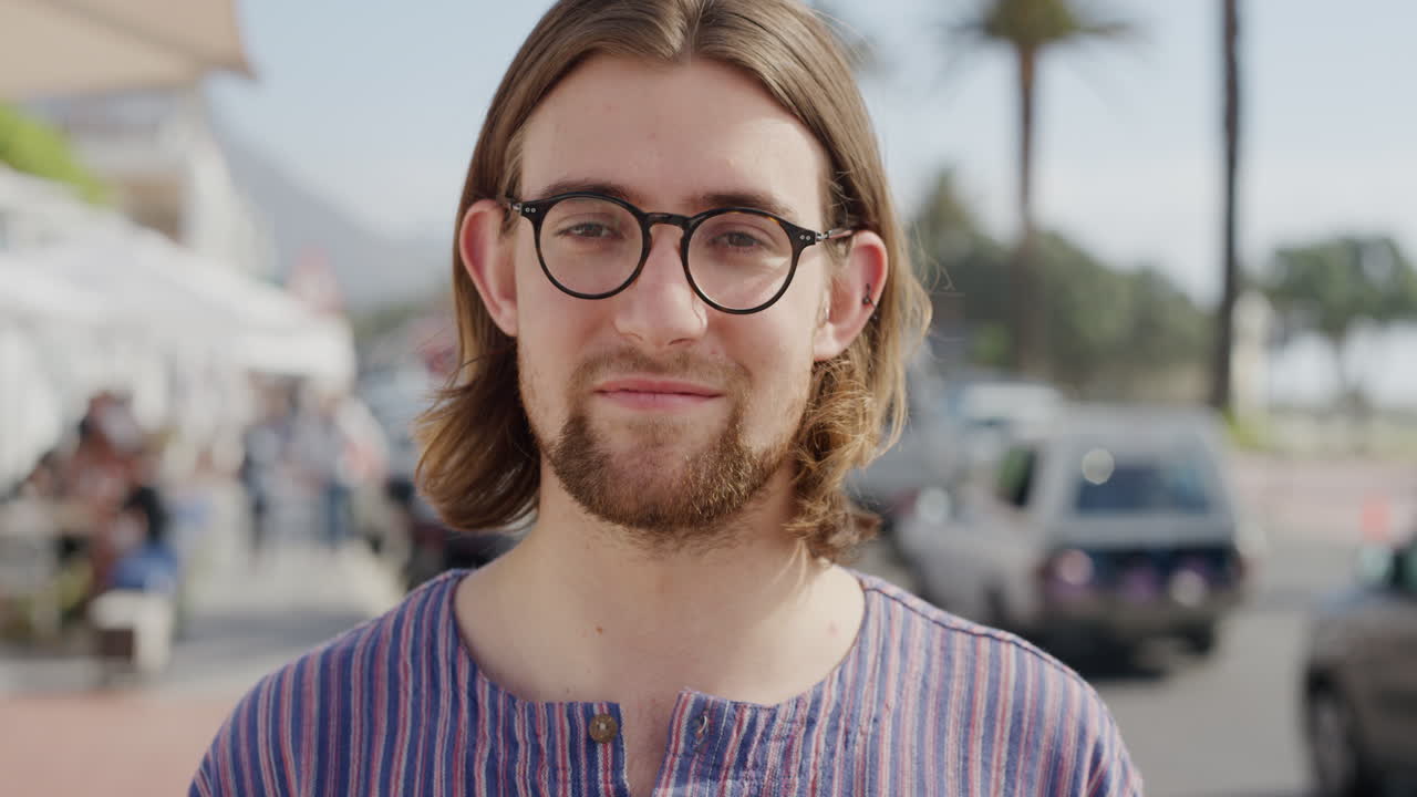ritratto di un simpatico giovane geek che indossa occhiali sorridente che si gode le vacanze estive all'aperto soleggiato che sembra calmo sulla spiaggia urbana affollata serie di persone reali
