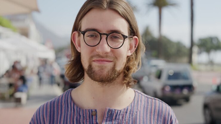 ritratto di un simpatico giovane geek che indossa occhiali sorridente che si gode le vacanze estive all'aperto soleggiato che sembra calmo sulla spiaggia urbana affollata serie di persone reali