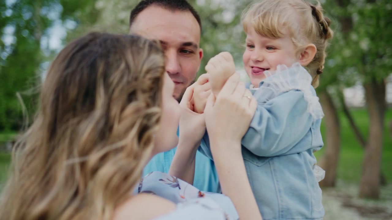 Glückliche Familie spielt draußen. Der Vater lächelt, während er seine Tochter hält. Die Mutter gesellt sich dazu, alle lachen. Ein warmer, fröhlicher Moment, der familiäre Liebe einfängt.