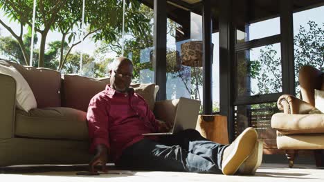 african american senior man sitting on living room floor working, holding paperwork and using laptop