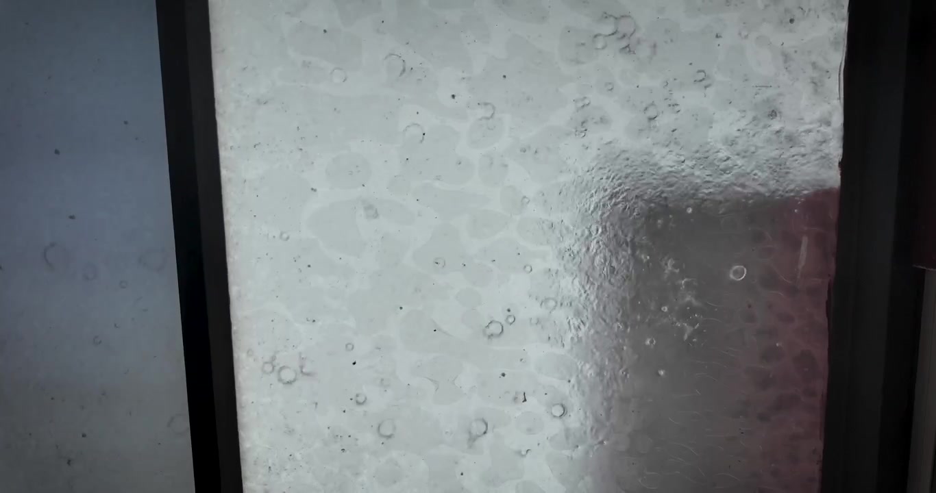 Heavy rainfall covers a window with moving droplets and flowing water while the urban landscape of Mexico City appears blurred in the background during a storm.