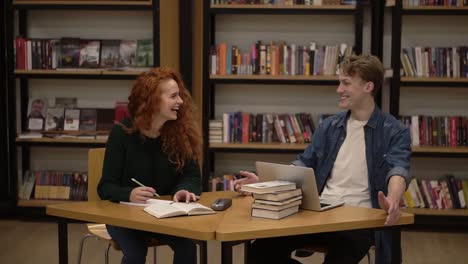 Young-Male-And-Female-Students-Talking-Using-Laptop-Explaining-Course-Work-Study-Together-In-Library