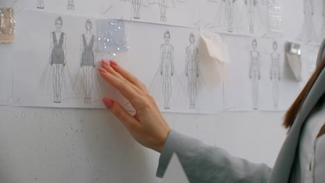 female fashion designer looking at drawings and sketches that are pinned to the wall behind her desk. studio is sunny. personal computer colorful fabrics sewing items are visible.