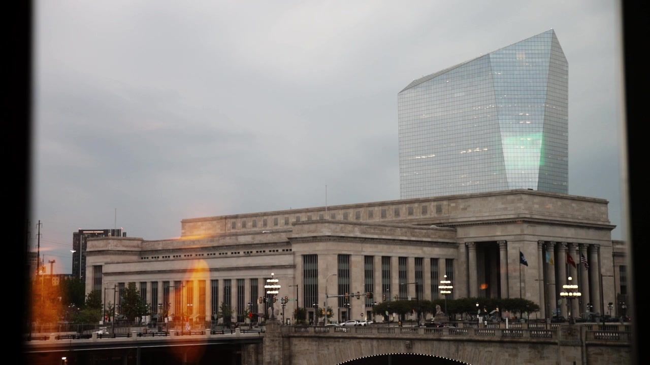 Vista da janela de um trem em movimento mostrando a histórica estação 30th Street e o moderno arranha-céu Cira Centre South, com o trânsito fluindo na rua abaixo sob um céu nublado.