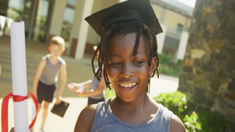 Video-De-Un-Feliz-Niño-Afroamericano-Con-Sombrero-De-Graduación-Y-Diploma-Frente-A-La-Escuela