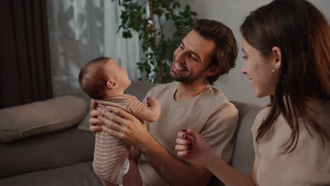 feliz joven moreno con paja el padre de una niña pequeña sostiene a su hija en sus brazos y se sienta en el sofá con su joven esposa en un apartamento moderno