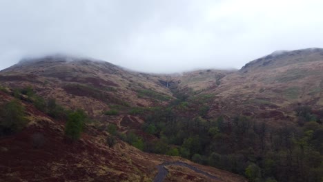 Aerial drone footage flying over green and brown colored woody Scotland landscape that is in an area with hills and mountains during a misty and foggy day when clouds are low and between the mountains