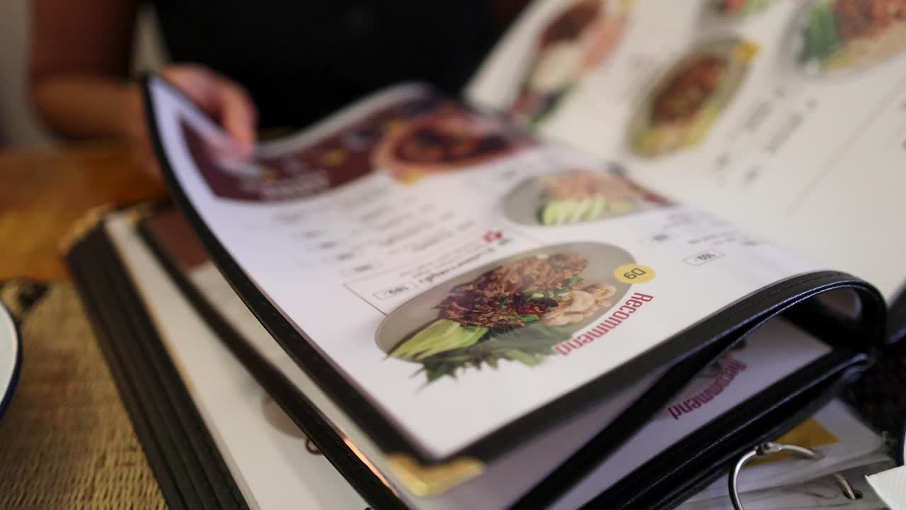 A person flips through a laminated menu featuring Northern Thai cuisine at a wooden restaurant table, under soft natural lighting with shallow depth of field