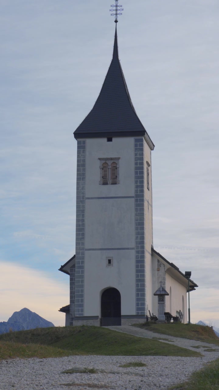 Un video verticale mostra una pittoresca chiesa in cima a una collina sullo sfondo di montagne, catturato con luce diurna soffusa e composizione stabile per uso mobile e sui social media.