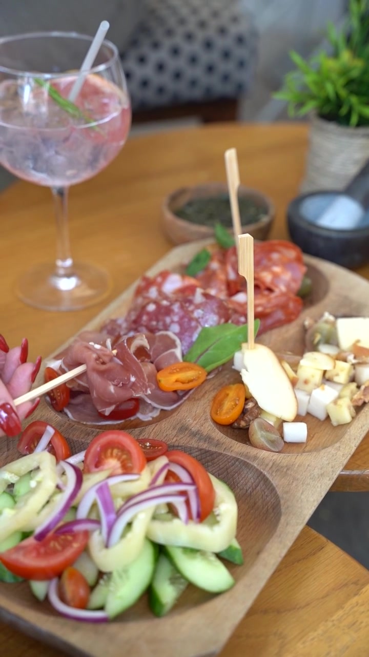 Una tabla de embutidos y quesos con una variedad de carnes curadas, quesos en lonchas y ensalada fresca con pepinos, tomates y cebolla roja. Se ven manos cogiendo los alimentos