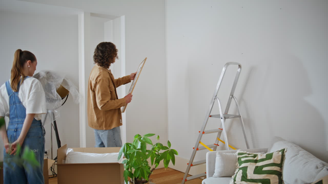 Girlfriend watching boyfriend decorating living room hanging artwork on wall